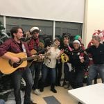 Officers in festive holiday gear join a guitarist and sing next to a Christmas tree in a community center space.