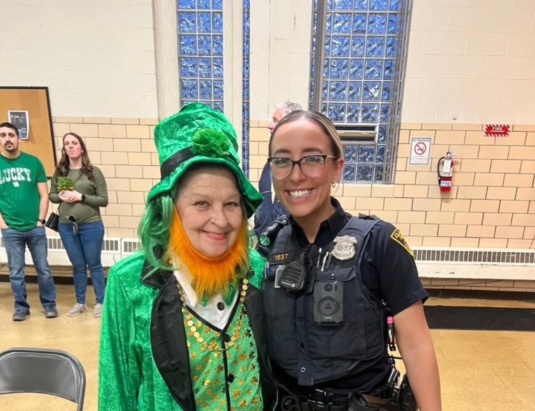 Officer Rodriguez poses with a smiling community member dressed in a bright green leprechaun costume at the Edna House event. Festive decorations and attendees in green attire are visible in the background.