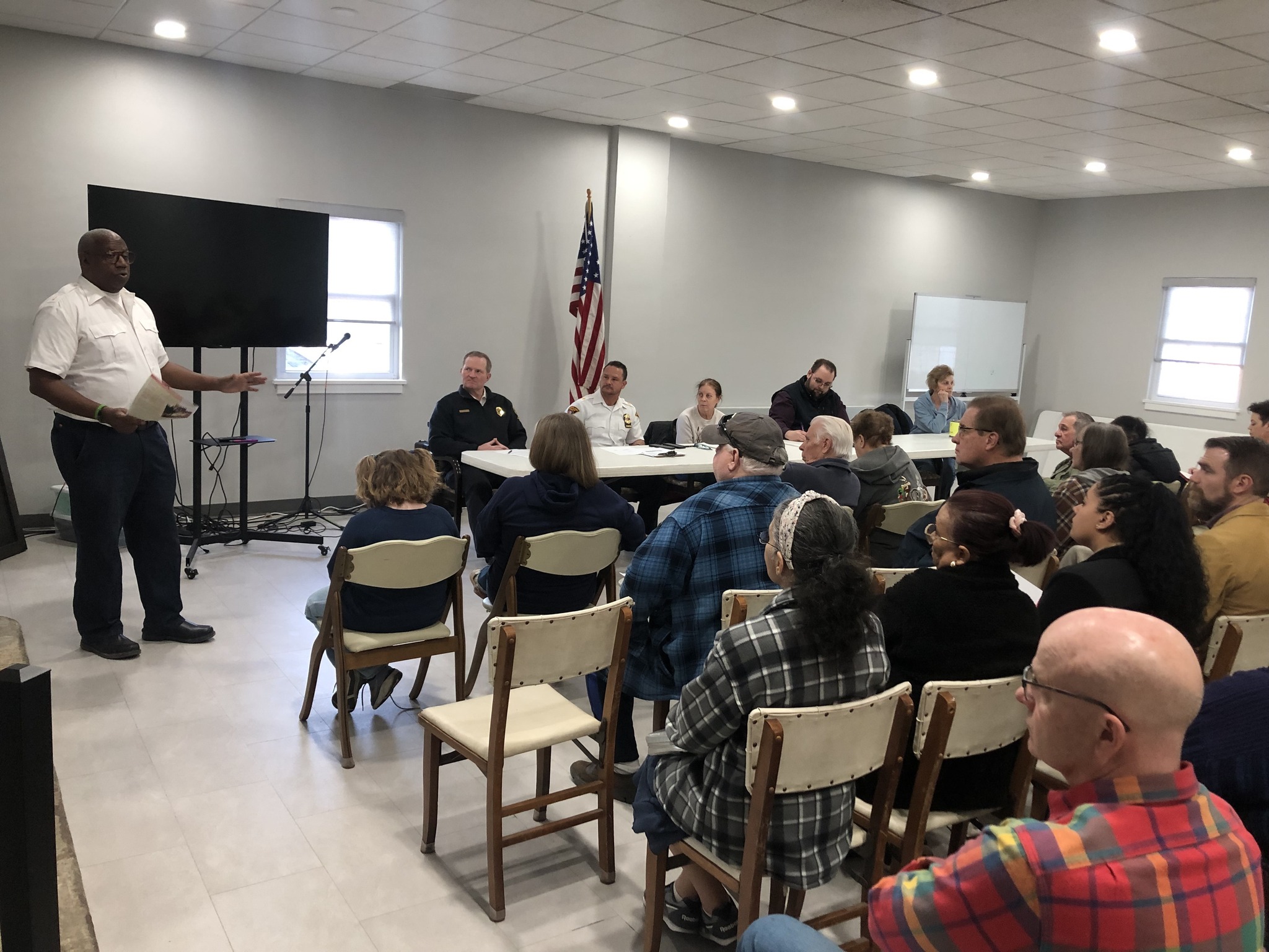 Residents and community members attend the Second District Community Meeting, listening to a speaker at the front of the room. Police officers sit at a table as part of the discussion, with an American flag and a large screen in the background.