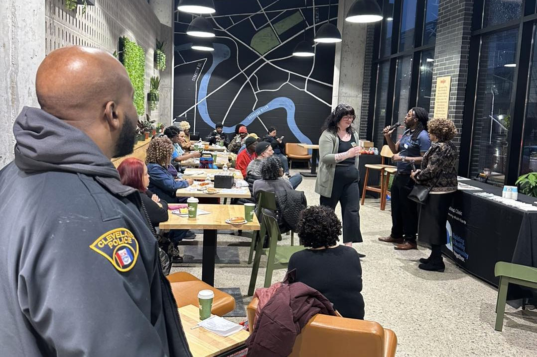 A Cleveland police officer observes a community gathering at Meijer Fairfax Market. Residents and stakeholders sit at tables while speakers lead a discussion at the front of the room. The space features modern décor with a large mural and hanging lights.