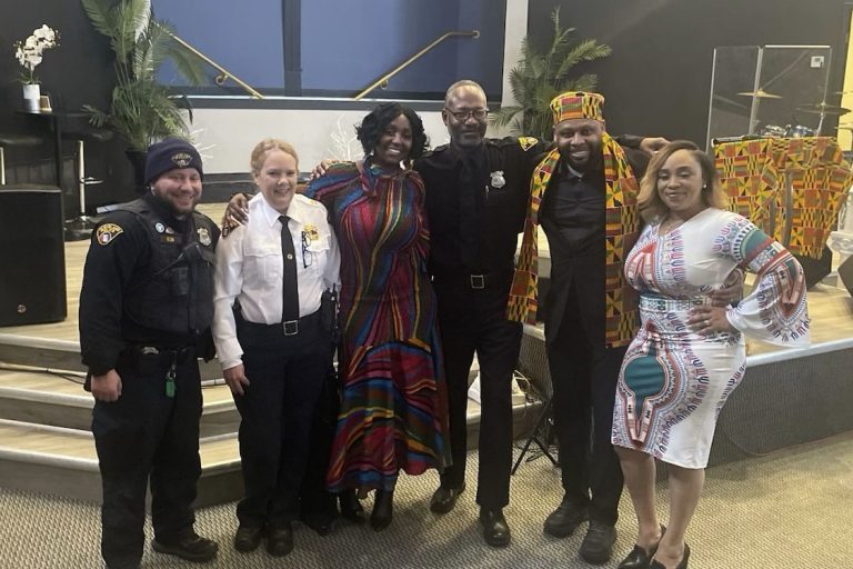 A group photo of Cleveland police officers and community members at Covenant Community Church's Black History Program. The group stands together smiling in front of a stage decorated with colorful African-inspired fabric and greenery.