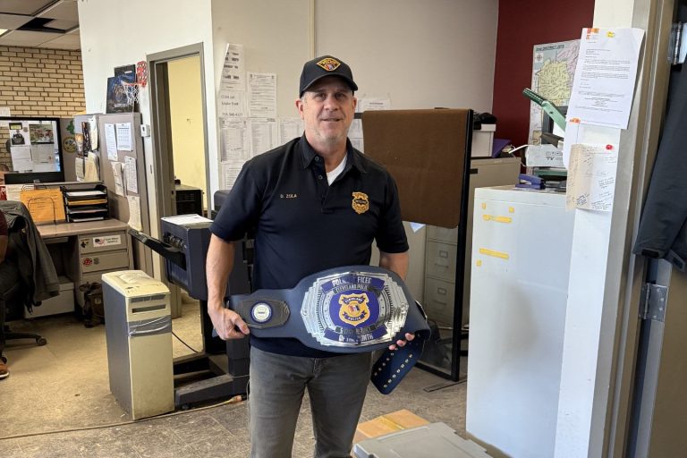 Detective Zola stands in an office holding an oversized championship-style belt recognizing him as Second District’s Officer of the Month.