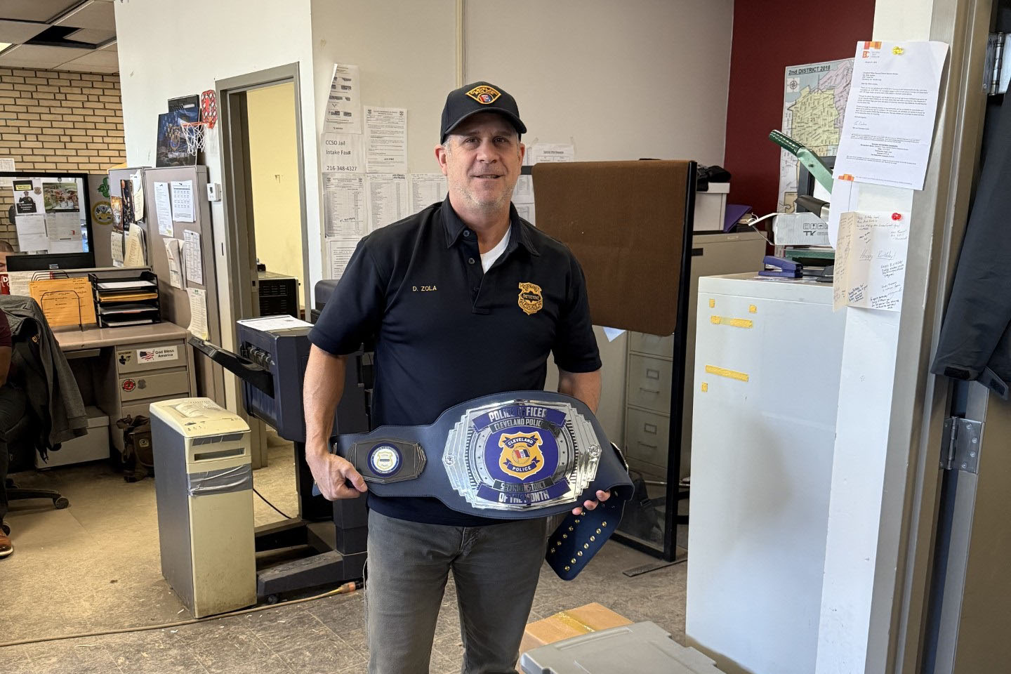 Detective Zola stands in an office holding an oversized championship-style belt recognizing him as Second District’s Officer of the Month.