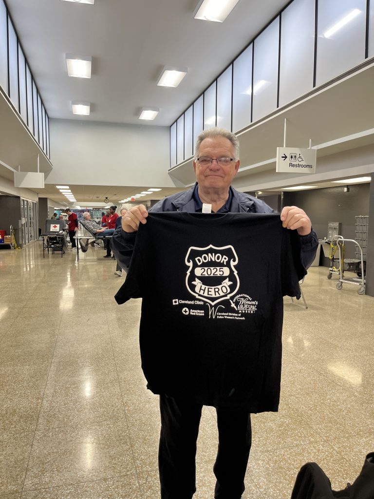 Bob Guttu holding a 2025 "Donor Hero" t-shirt, standing in the center of the donation area.