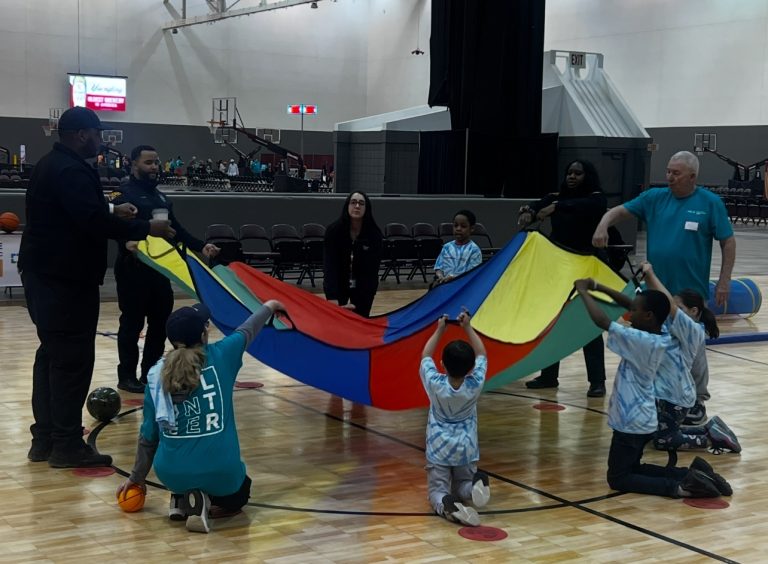 Kids, officers, and volunteers raise a colorful parachute during a court activity.