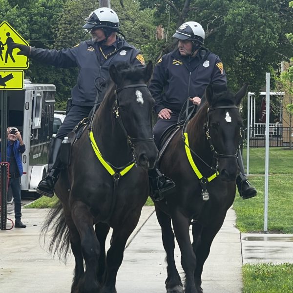Two Mounted Unit officers ride side-by-side down the sidewalk as students watch.