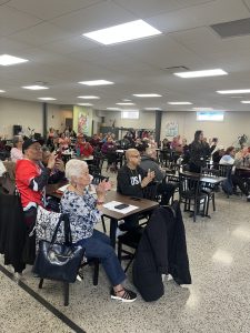 A large group of seniors seated at tables applauds during the Community Partner of the Month presentation.