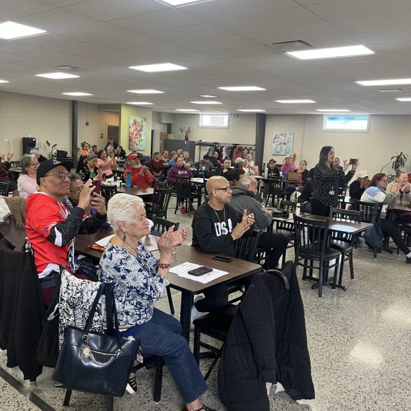 A large group of seniors seated at tables applauds during the Community Partner of the Month presentation.