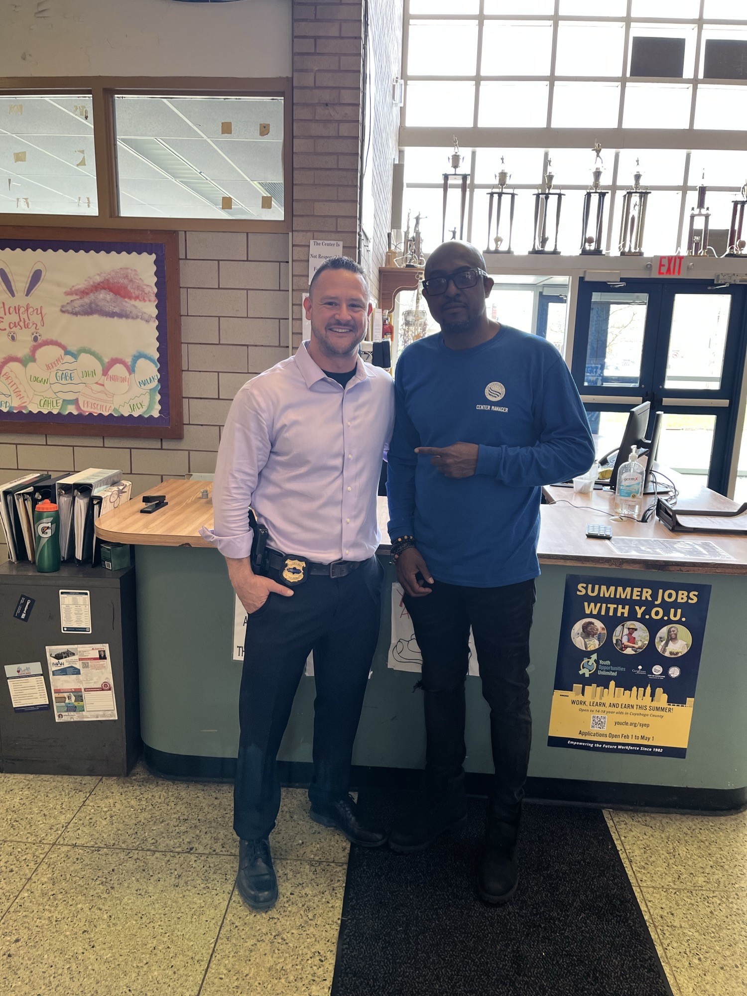 Estabrook Rec Center Commander Maffo-Judd stands with Estabrook Rec Center Director Jack Nichols near the front desk, with trophies and a youth job flyer in the background.