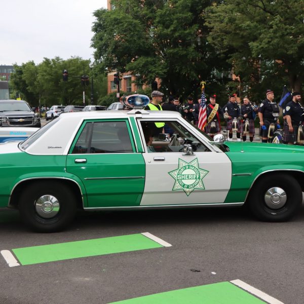 Vintage sheriff’s patrol car drives past as out-of-town police pipe bands line up in the background