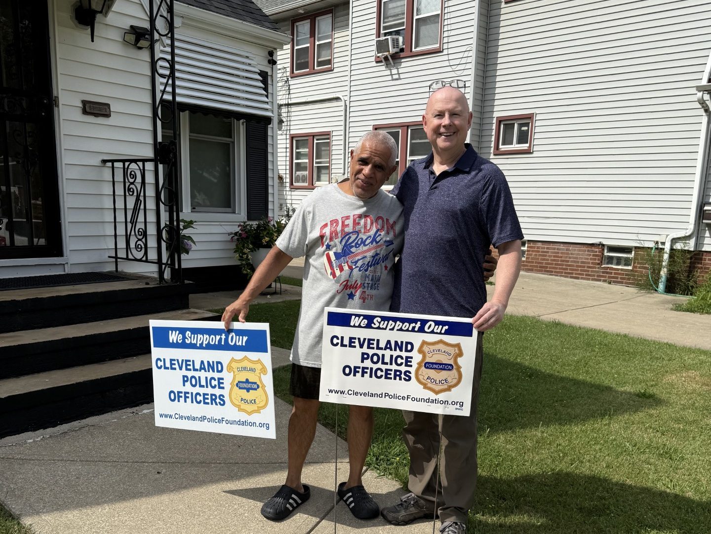 Support Our Police Yard Signs Mr. Pena stands outside his home holding a “Support Our Police Officers” yard sign alongside a supporter, both smiling on the sidewalk.